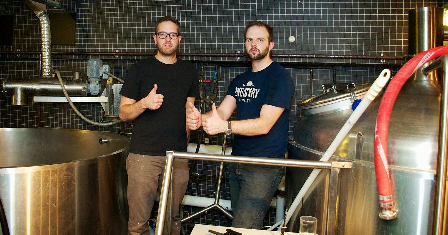 two brewers standing on a platform next to a mash tun and kettle